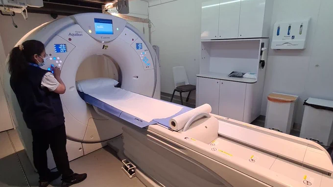 A radiographer is standing next to a CT scanner pushing a button on the machine. 