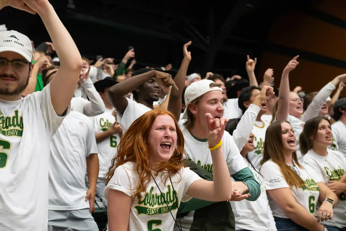 Colorado State University's Moby Arena is filled with enthusiastic basketball fans. The crowd, dressed in white shirts, is cheering passionately and raising their hands to make the CSU Rams hand sign.
