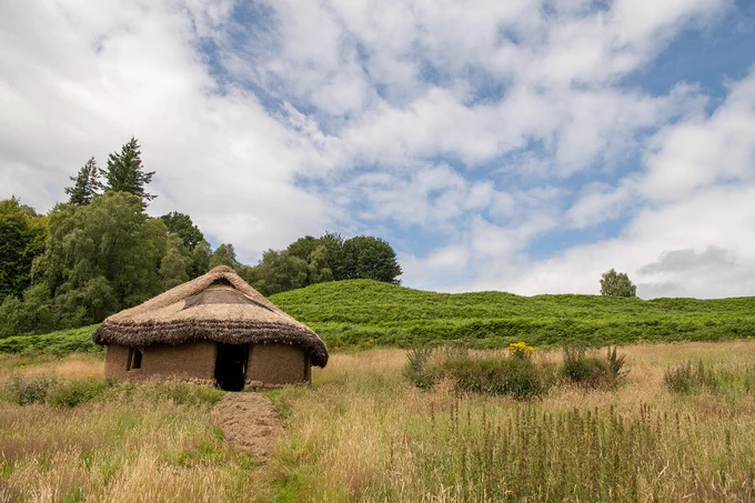 A small, thatched roof hut in the countryside. The hut is made from earth and has a large, rectangular opening for a door.