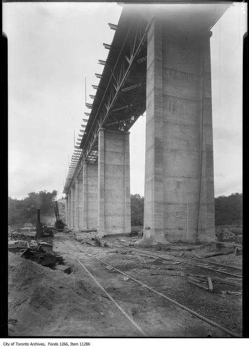 Black and white photo of the concrete pillars of the Leaside Bridge, seen from the ground. High above, the steel girders are in place, but only some of the road surface has been laid down. Fonds 1266, item 11286.