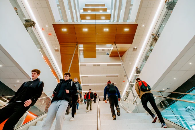 Students walk up and down a long straight staircase that separates the first and second floor before the stairs wind up the east side of the atrium.