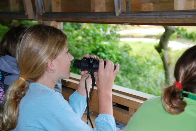A teenage girl with brown hair in a ponytail. She is holding binoculars and looking out from a bird hide.