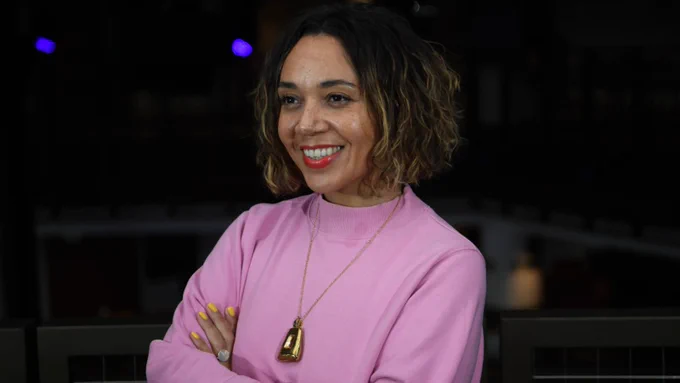 A photo of Raidene, a brown-skinned female, aged early forties, with wavy brown shoulder-length hair, wearing a pink jumper, looking away from the camera, smiling
