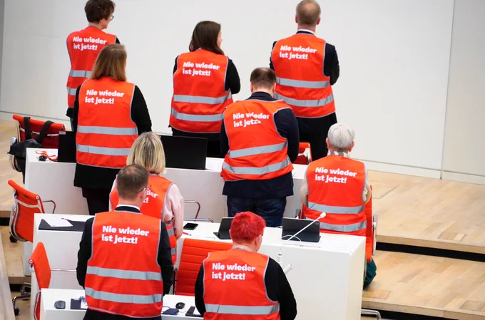 Ein Foto unserer Abgeordneten im Plenarsaal, mit dem Rücken zur Kamera gedreht. Sie tragen rote Warnwesten mit der Aufschrift "Nie wieder ist jetzt!"