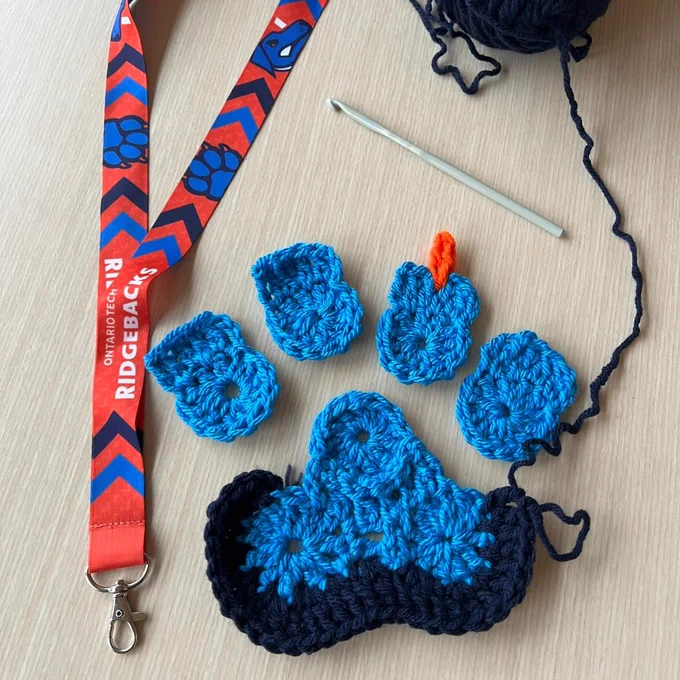 A Ridgebacks lanyard sits on the table next to a crochet project in process that in the shape of the Ridgebacks paw logo in dark blue, light blue and orange. A dark blue skein of yarn and a hook are at the top of the frame. 