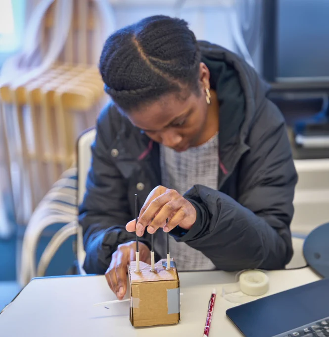 Young female student is working on a prototype made of cardboard and plastic pegs. Black coat, indoor. 