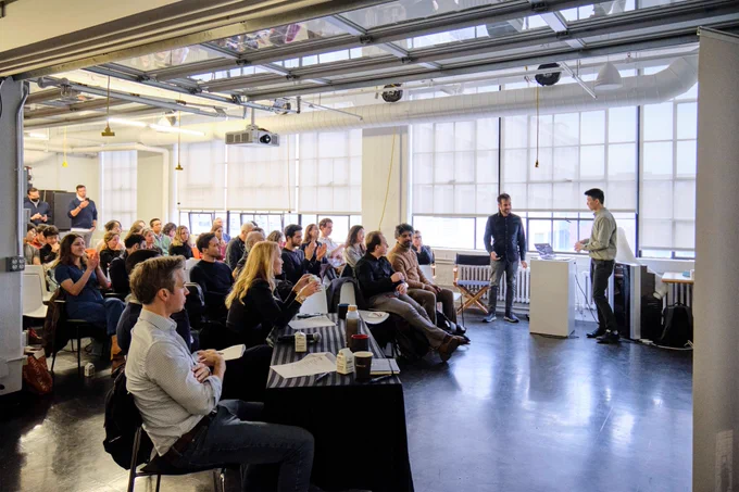 An audience is gathered in a large room, attending a startup pitch. One man stands, presenting. Indoors. 