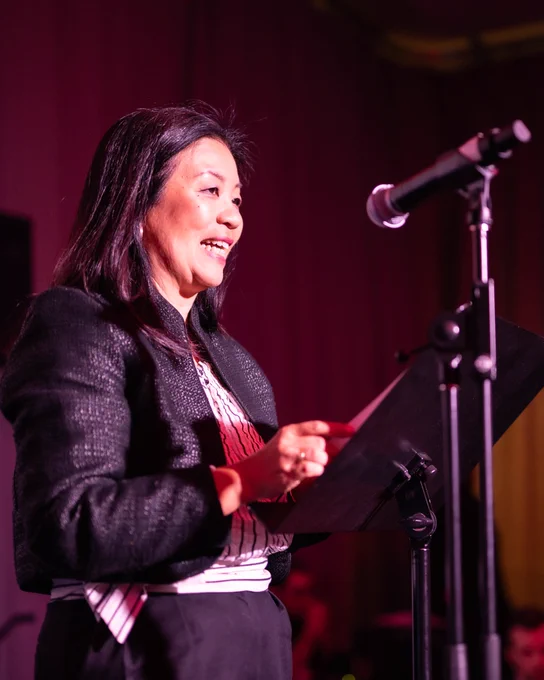 A photograph of TIFF Chief Programming Officer Anita Lee speaking on stage at an event, with a microphone in the foreground  and a podium in front of her.