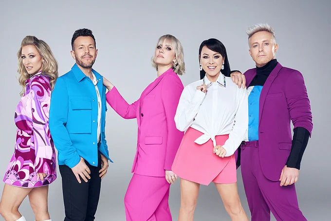 A photo showing five performers in vivid outfits, posing against a grey background