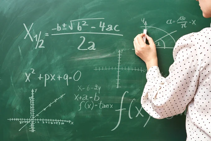 A math educator writing on a green chalkboard with white chalk.