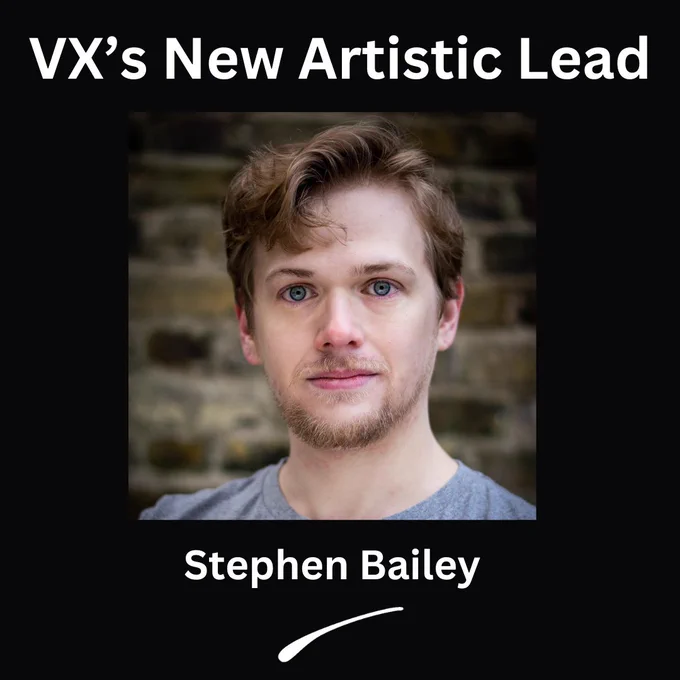 A white man with blue eyes, blond hair, glasses and facial hair. They are wearing a grey t-shirt. White letters read: VX's new Artistic Lead. Stephen Bailey.