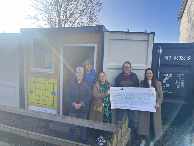 A group of 5 people hold a large cheque in front of the Cadog’s Corner shop.