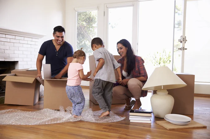 Photo of family moving into their new home and unpacking boxes. Small children stepping on bubble wrap while their parents watch smiling.