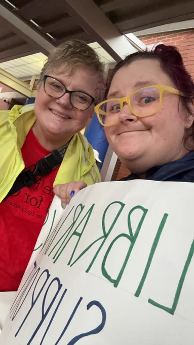 2 white women that are drenched with rain stand together holding a protest sign that reads 'Librarians support classified staff'