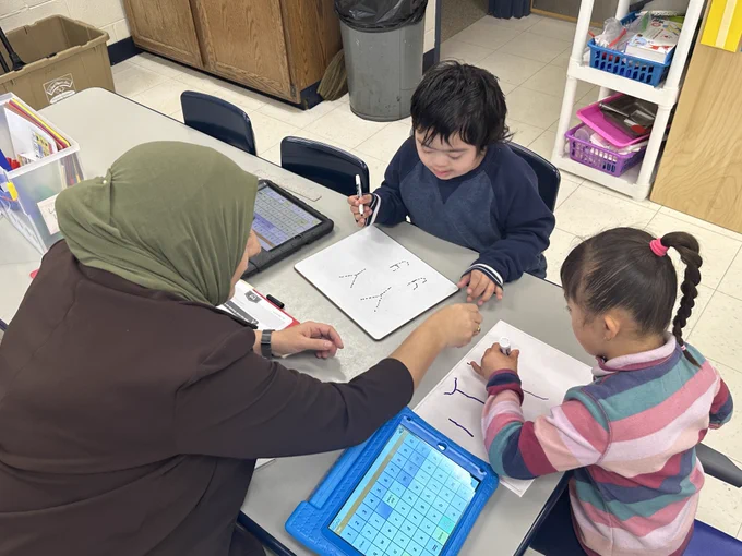 Image of a teacher sitting at a table with two students working on letters with dry erase boards
