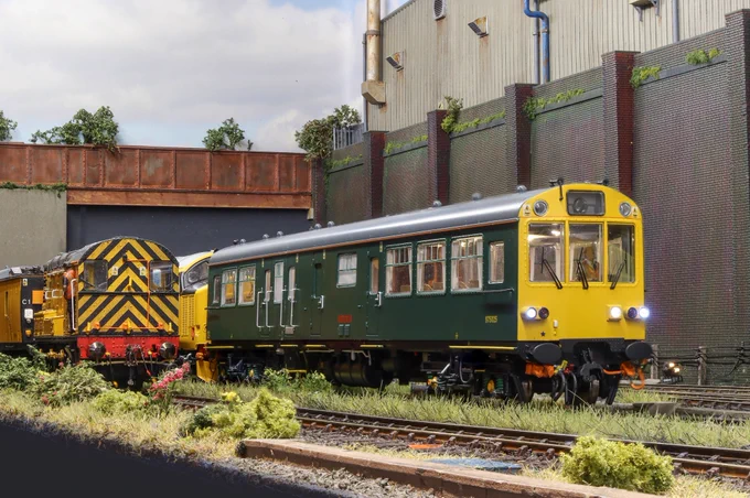 A model railway diorama of an industrial scene. A green inspector's saloon is sat in a railway yard next to a Class 08 shunter.