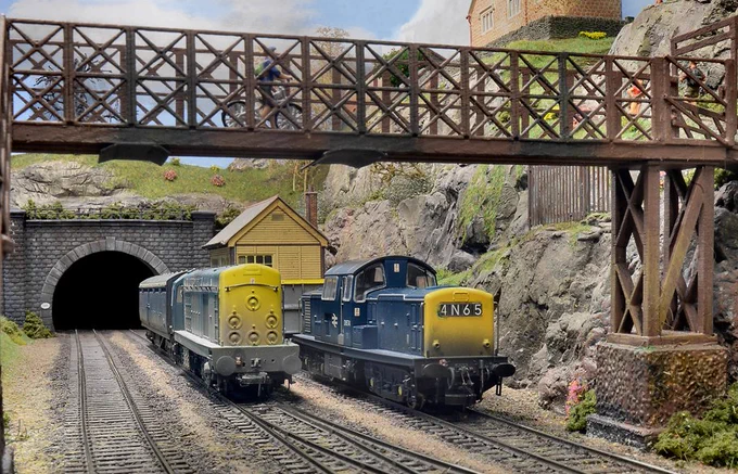 A model railway diorama scene. A Class 20 and Class 14 locomotive sit on the track in a cutting with a metal footbridge above and a tunnel mouth behind.