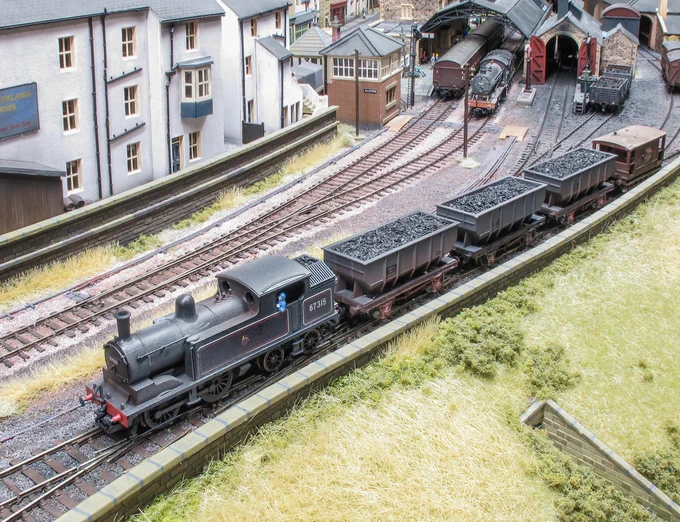 A model railway diorama showing a British Railways steam tank engine hauling wagons filled with coal out of a station.