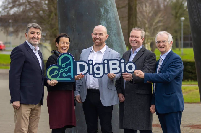 A group of five people stand in front of a large sculpture holding a sign of a heart with the word 'DigiBio' written beside it. 