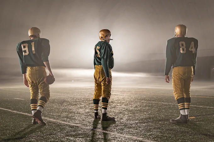 Three actors playing Edmonton football players on a field at night with bright lights in background.