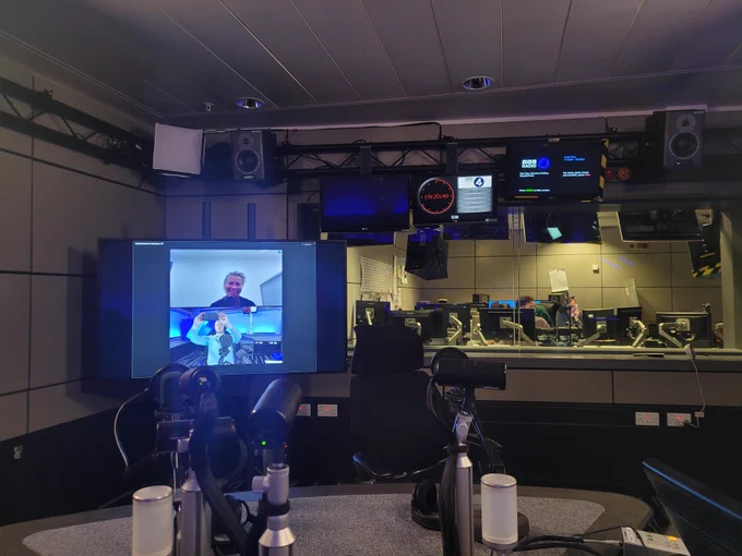 BBC Radio studio, with microphones, desks and gallery of producers, with a big TV screen with Sam White on it.