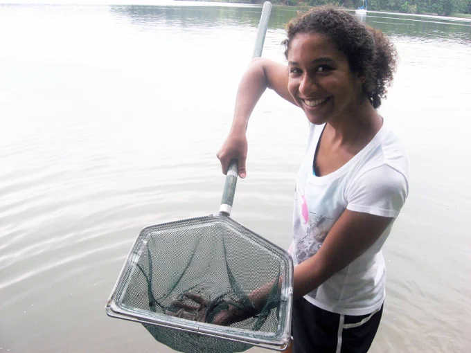 Young intern stands in a river, holding a net with a long handle and smiling.