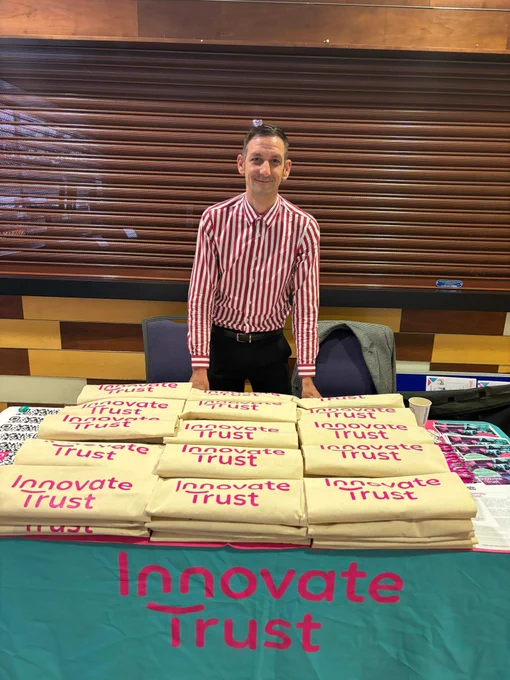 Operations Manager, Mikey, smiling while leaning on Innovate Trust's stall at Cardiff Job Fair. The stall is filled with posters and Innovate Trust tote bags
