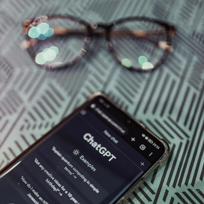 Ein Handy und eine Brille liegen auf einem Tisch. Auf dem Bildschirm ist ChatGPT geöffnet.
