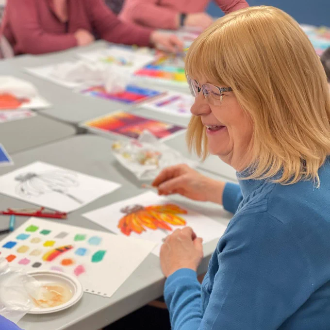 A woman sits at a craft table and enjoys creating an oil pastel flower while turned and laughing with her friend at the table.