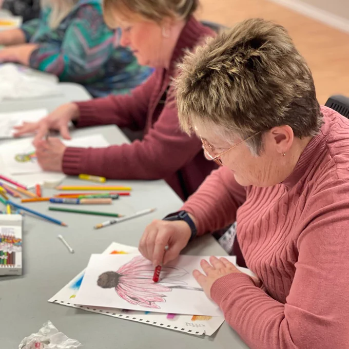 A woman sits at a crafting table creating a pink-hued flower using oil pastels.