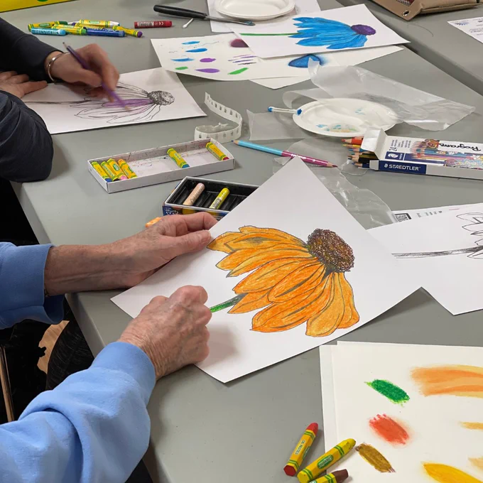 A side-view of two hands engaged in drawing an orange and yellow flower with oil pastels.