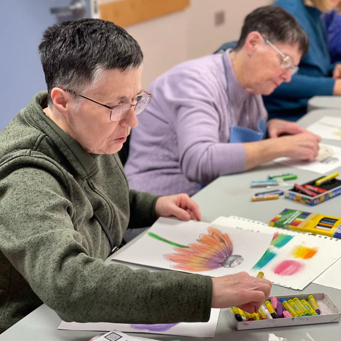 A woman at a crafting table creating a purple and orange flower using oil pastels.