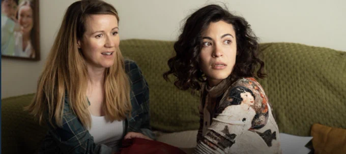 Two women sat on the green sofa looking out to the window. The woman on left is looking with no focus, has long blonde hair and wearing a checked blue shirt with a white top. The woman on the left is looking at something has short dark curly hair and wearing a patterned brown and cream top. There is a family picture in the background on a cream wall.



