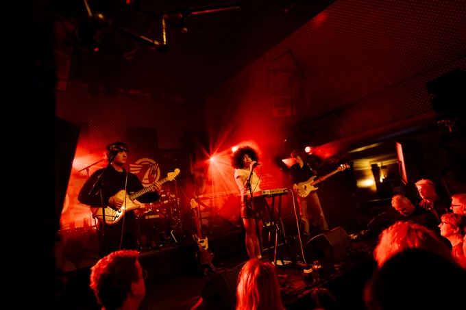 Band performing on a stage with people watching them, the lighting is red. The band consists of a singer, a guitar player, a bass player, and a drummer.