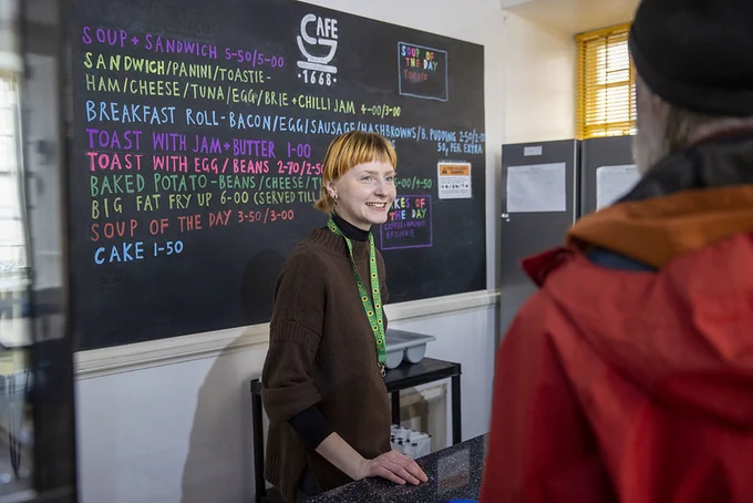 A woman stands smiling behind a counter, a lunch menu written on the wall behind her