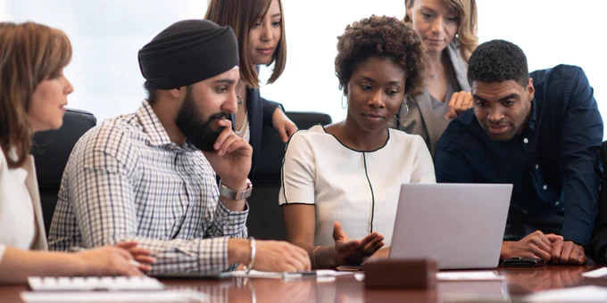 A group of diverse people gathered around a laptop in discussion.