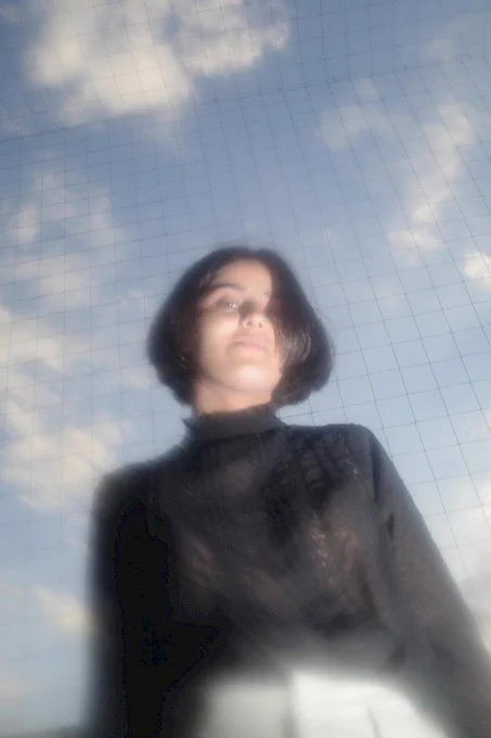 A portrait of the DJ babyshon. She has a black bob and is wearing a black jumper against a background of clouds and blue sky