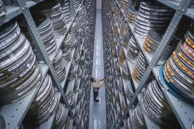 A person standing in between two large shelfing units, with film reels stacked up on the shelves.