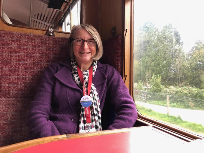 
This is an image of Hayley Buscombe, Community Engagement Lead, for Devon Carers. She is on a train looking very happy. She is wearing a purple coat and a black and white checkered scarf.
