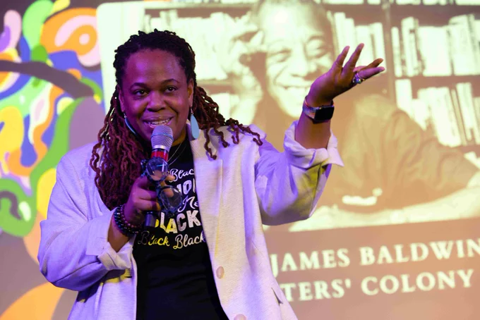 A smiling person speaks with a microphone and gestures with the other hand.