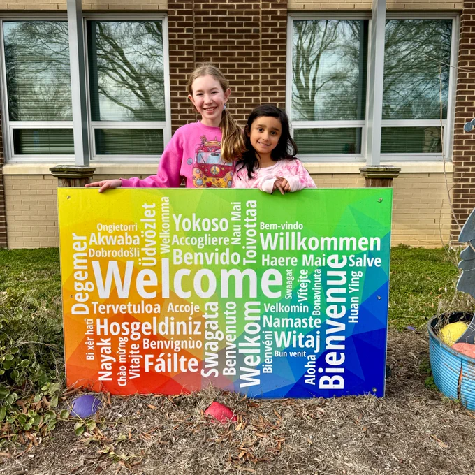 Two students standing behind a colorful sign that has the word "welcome" written in multiple languages.