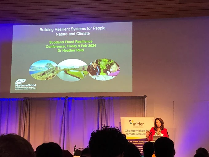 A woman in a red jacket stands on a stage delivering a speech watch by an audience of people. The slide on the wall behind her has a block of text which reads:

Building Resilient Systems for People, Nature and Climate
Scotland Flood Resilience Conference, Friday 9 Feb 2024
Dr Heather Reid
NatureScot

There are three oval shaped interlinked photos showing a tree growing on a loch shore, a watercourse running past some buildings, and two people taking photographs with a camera outside.