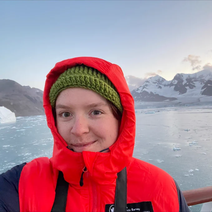 Emily Broadwell, a University of Bristol PhD student, smiles in front of sheet ice and snow-capped mountains. She is dressed for very cold weather.