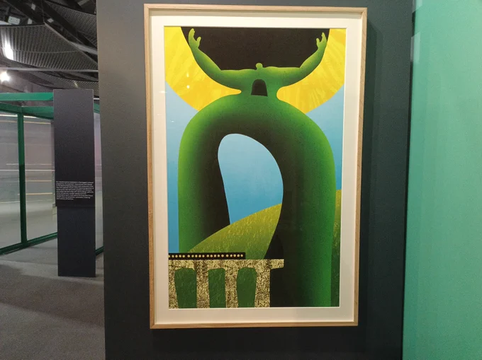 A large poster print framed on a gallery wall. The print is an abstract image and has similar shapes and colours to a sunflower. There is a human figure in green holding up a yellow and black orb. There is also a train track in the bottom left hand side. 