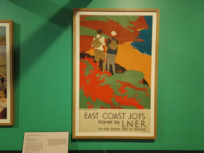 A poster print on a green gallery wall. The poster is an illustration of two people hiking on a cliff. The text says: 'East Coast Joys travel by L.N.E.R. To the drier side of Britain'.