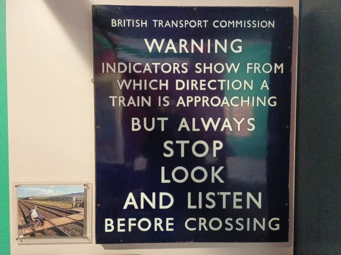 A metal sign and image on gallery wall. The metal sign is laquered blue and has the text: 'British Transport Commission. Warning. Indicators show from which direction a train is approaching but always stop look and listen before crossing'. The image is of a person in a wheelchair crossing a path over the traintracks.