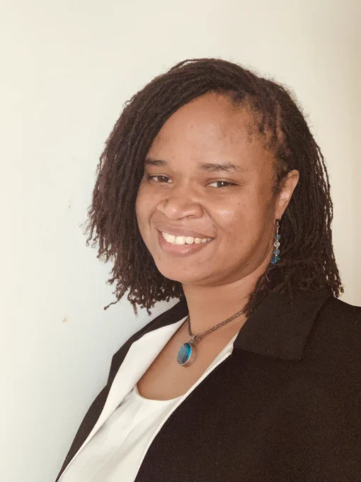 Syreeta, a black woman with shoulder length dark brown Sisterlocs parted to the side and dark brown eyes smiling at the camera with an off-white background. She is wearing blue earrings, a circular blue necklace, a white shirt, and a business jacket with a white collar