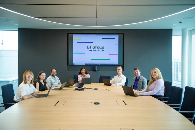 7 people sitting around a table with laptops in front of them. They are smiling and looking at the camera. The BT Group logo is on display behind them.