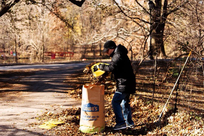 A side view of a man in a black raking leaves into a large brown paper bag. There is a lot of foliage around him and leaves on the ground.