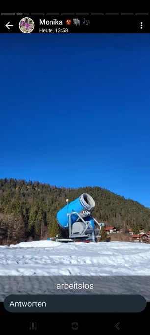 Eine Schneekanone steht im Landkreis Garmisch-Partenkirchen ohne Betätigung auf etwas Schnee herum. Im Hintergrund ein Hügel, Häuser. Alles sieht eher nach Frühling aus 
